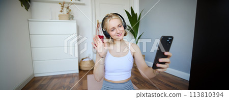 Portrait of young woman with smartphone and headphones, waving hand at mobile phone camera, live streaming during workout Portrait of young woman with smartphone and headphones, waving hand at mobile phone camera, live streaming during workout 113810394