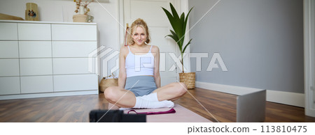 Portrait of young fitness instructor, vlogger showing exercises on camera, recording herself, sitting on mat with laptop, doing workout, explaining yoga movements to followers 113810475
