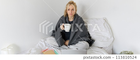 Portrait of woman catching a flu, sneezing, feeling sick, sitting on bed with laptop and working on remote while caught a cold 113810524