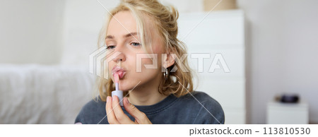 Close up portrait of beautiful blond woman applies lip gloss on her lips, puts on makeup, gets ready for night out, using beauty products, looks in mirror 113810530