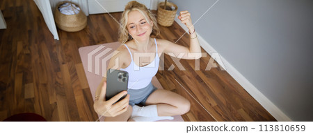 Portrait of young sporty woman taking selfie during workout training, content creator takes photos, shows muscles on camera, flexing biceps 113810659