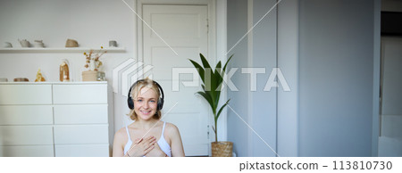 Portrait of woman feeling relaxed and in peace after meditation or yoga training at home, holding hands on chest, wearing headphones, smiling at camera Portrait of woman feeling relaxed and in peace after meditation or yoga training at home, holding hands on chest, wearing headphones, smiling at camera 113810730