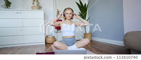 Portrait of young fitness girl, woman workout at home, listens to fitness instructor in wireless headphones, follows exercises on laptop 113810740