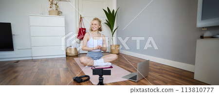 Portrait of young fitness blogger, woman showing exercises to her followers, recording video on digital camera, doing workout training session, home yoga, sitting in front of laptop on rubber mat 113810777