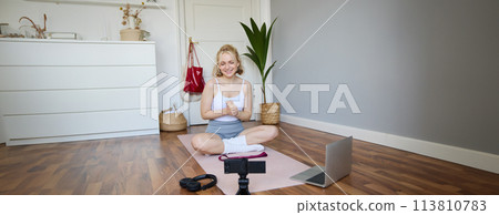 Portrait of young fitness blogger, woman showing exercises to her followers, recording video on digital camera, doing workout training session, home yoga, sitting in front of laptop on rubber mat Portrait of young fitness blogger, woman showing exercises to her followers, recording video on digital camera, doing workout training session, home yoga, sitting in front of laptop on rubber mat 113810783