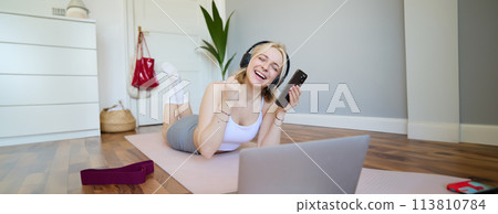 Portrait of woman lying on rubber yoga mat in room, wearing headphones, listens to music, using laptop and smartphone Portrait of woman lying on rubber yoga mat in room, wearing headphones, listens to music, using laptop and smartphone 113810784