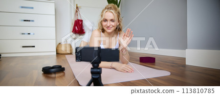 Portrait of smiling fitness instructor, social media content creator recording a vlog about sport, using rubber yoga mat, waving hand and looking at digital video camera 113810785