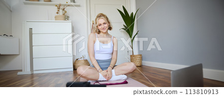 Portrait of young fitness instructor, vlogger showing exercises on camera, recording herself, sitting on mat with laptop, doing workout, explaining yoga movements to followers 113810913