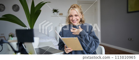 Portrait of smiling young cute woman, holding notebook, girl with laptop and planner in hands, sits in room, looks at digital camera, creates lifestyle vlog, talks to audience, reads script Portrait of smiling young cute woman, holding notebook, girl with laptop and planner in hands, sits in room, looks at digital camera, creates lifestyle vlog, talks to audience, reads script 113810965