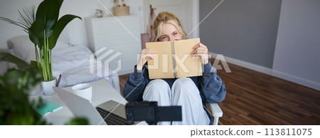 Portrait of cute, smiling young social media content creator, girl records video on digital camera and stabiliser, holds notebook, talks to audience, vlogging in her room 113811075