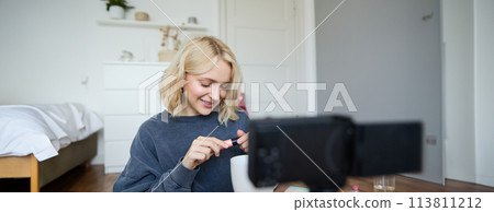Close up portrait of smiling young woman with video camera in her room, showing mascara, reviewing beauty products for social media account. Beauty blogger creates vlog about makeup 113811212