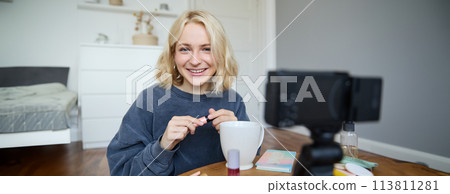 Portrait of beautiful, smiling blond woman, girl recording video of her makeup tutorial for social media, vlogger sitting on floor in her room, using stabiliser to create content, reviewing mascara 113811281