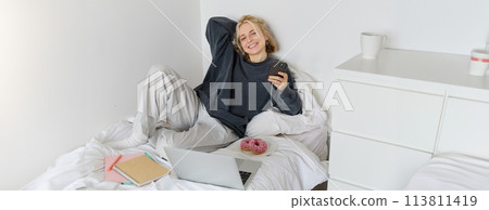 Portrait of young beautiful blond woman, feeling lazy at home, lying in bed with cup of tea, doughnut and laptop, studying remotely or watching movie online Portrait of young beautiful blond woman, feeling lazy at home, lying in bed with cup of tea, doughnut and laptop, studying remotely or watching movie online 113811419