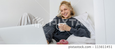 Portrait of candid, happy young woman lying in bed, looking at laptop screen, holding cup of tea and eating doughnut, staying at home on weekend, spending quite time alone 113811451