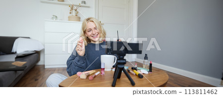 Image of cheerful, beautiful young lifestyle blogger, woman sitting on floor and recording video about makeup, holding mascara, making lifestyle content for her social media account and followers 113811462