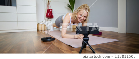 Portrait of young sporty vlogger, fitness instructor standing in plank on rubber yoga mat, recording video of herself doing workout at home 113811503
