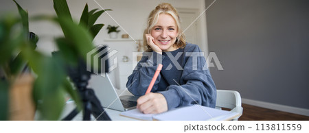 Cute smiling girl sits in a room, writes down notes, doing homework, records video of herself on digital camera, creates content for vlog, lifestyle blogger doing daily routine episode 113811559
