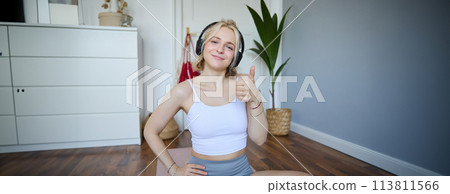 Portrait of beautiful, healthy woman in headphones, showing thumbs up, sitting on rubber mat, recommending yoga online training video, pleased with exercises for home training Portrait of beautiful, healthy woman in headphones, showing thumbs up, sitting on rubber mat, recommending yoga online training video, pleased with exercises for home training 113811566