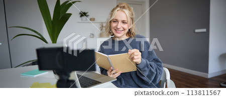 Portrait of young woman using digital camera and laptop, studying, connect to online live stream, records video for vlog, creating content, holding notebook, sitting in room 113811567