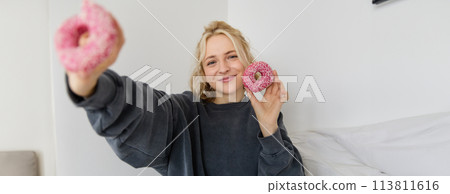 Portrait of beautiful smiling blond woman, showing two pink doughnuts at camera, eating delicious food 113811616