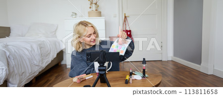 Portrait of smiling blond woman, teenage girl records a vlog on video camera, vlogger shows eyeshadows, tutorial how to put makeup on, sits in her room on floor near coffee table Portrait of smiling blond woman, teenage girl records a vlog on video camera, vlogger shows eyeshadows, tutorial how to put makeup on, sits in her room on floor near coffee table 113811658