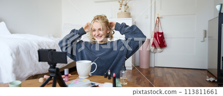 Portrait of cute blond girl, lifestyle blogger sits on floor with video camera and stabiliser, shows how to do hairstyle and makeup, does casual vlogging for social media, sits in room Portrait of cute blond girl, lifestyle blogger sits on floor with video camera and stabiliser, shows how to do hairstyle and makeup, does casual vlogging for social media, sits in room 113811661