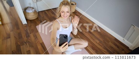 Portrait of fit, sporty young fitness instructor, woman gives online workout sessions to clients, waves hand at smartphone, sits on yoga mat and shows exercises 113811673