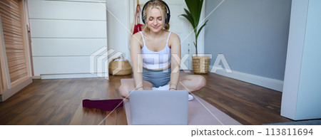 Portrait of young sporty woman following online video instructions during fitness workout, using laptop, sitting on yoga mat Portrait of young sporty woman following online video instructions during fitness workout, using laptop, sitting on yoga mat 113811694