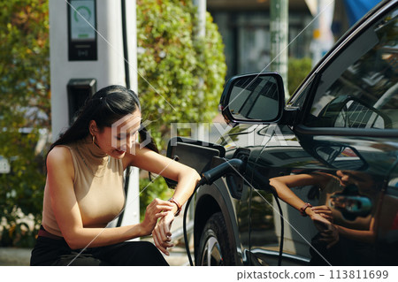 Smiling woman checking time on wristwatch when waiting for her electric car to fully charge Smiling woman checking time on wristwatch when waiting for her electric car to fully charge 113811699