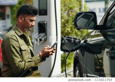 Frowning man texting when waiting for his electric car to charge Frowning man texting when waiting for his electric car to charge 113811703
