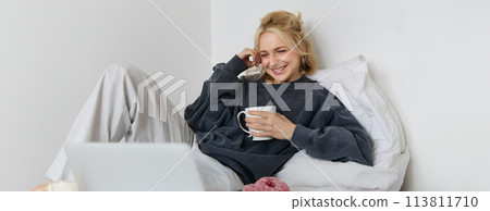 Portrait of candid, happy young woman lying in bed, looking at laptop screen, holding cup of tea and eating doughnut, staying at home on weekend, spending quite time alone Portrait of candid, happy young woman lying in bed, looking at laptop screen, holding cup of tea and eating doughnut, staying at home on weekend, spending quite time alone 113811710