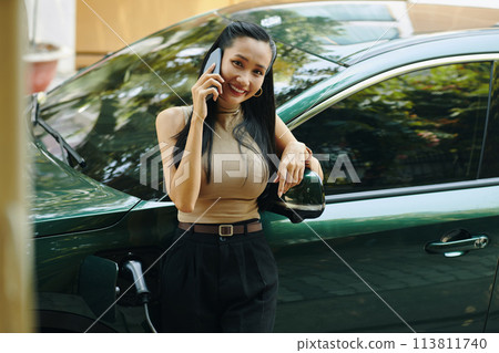 Smiling successful businesswoman standing next to electric car when talking on phone Smiling successful businesswoman standing next to electric car when talking on phone 113811740