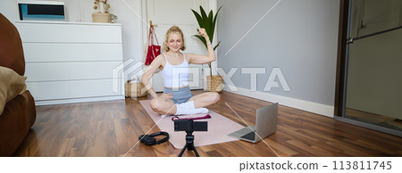 Portrait of young fitness instructor, vlogger showing exercises on camera, recording herself, sitting on mat with laptop, doing workout, explaining yoga movements to followers 113811745