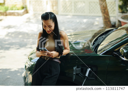 Smiling woman checking charge level of electric car battery in mobile app Smiling woman checking charge level of electric car battery in mobile app 113811747