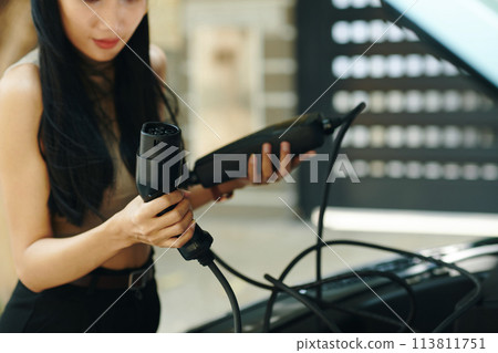 Cropped image of woman taking charging cable out of electric car trunk 113811751