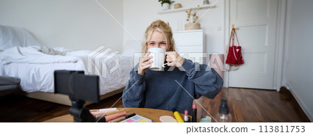 Image of young woman, makeup vlogger, sitting in bedroom with digital camera, drinking tea and talking, creating lifestyle video, social media content 113811753