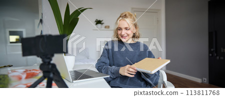 Young smiling blond woman, sits near laptop, uses digital camera to record video blog, creates lifestyle content for social media, records tutorial, shows notebook, reads her notes 113811768