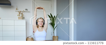Vertical shot of smiling, active and healthy woman workout at home, follows fitness instructions, watches video on laptop in headphones, stretches her arms Vertical shot of smiling, active and healthy woman workout at home, follows fitness instructions, watches video on laptop in headphones, stretches her arms 113811778