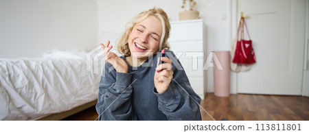 Portrait of smiling beautiful woman in her room, sitting and showing lipstick, recommending favourite beauty product, content maker recording a video of herself for social media blog Portrait of smiling beautiful woman in her room, sitting and showing lipstick, recommending favourite beauty product, content maker recording a video of herself for social media blog 113811801