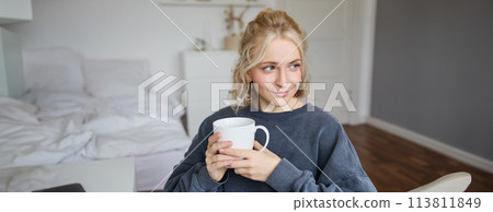 Portrait of smiling beautiful young woman, sitting in bedroom with cup of tea, resting at home alone, enjoys her weekend indoors 113811849