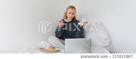 Portrait of sad, crying young woman, staying at home, sitting in bed with doughnut and comfort food, looking at something upsetting on laptop screen Portrait of sad, crying young woman, staying at home, sitting in bed with doughnut and comfort food, looking at something upsetting on laptop screen 113811867