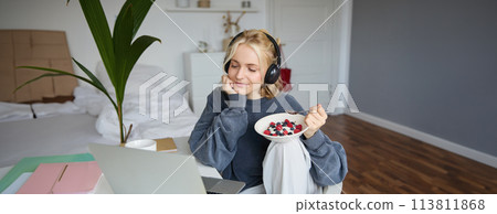 Image of happy woman sitting in a room, watching interesting tv show or movie on laptop, using screaming service, wearing headphones, eating dessert and drinking tea Image of happy woman sitting in a room, watching interesting tv show or movie on laptop, using screaming service, wearing headphones, eating dessert and drinking tea 113811868