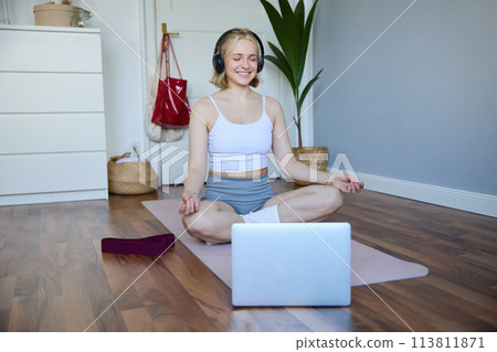 Portrait of young and relaxed woman using laptop and wireless headphones while meditating, practice yoga at home on rubber mat, sitting in asana Portrait of young and relaxed woman using laptop and wireless headphones while meditating, practice yoga at home on rubber mat, sitting in asana 113811871