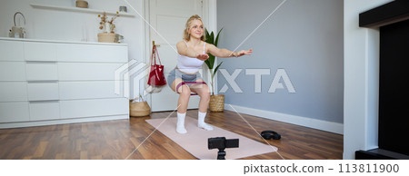 Portrait of woman, fitness instructor at home, recording video about workout, showing how to do leg exercises, squats with elastic resistance band, working out indoors on yoga mat Portrait of woman, fitness instructor at home, recording video about workout, showing how to do leg exercises, squats with elastic resistance band, working out indoors on yoga mat 113811900