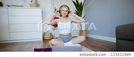 Portrait of fit and healthy young woman in wireless headphones, does workout, using online video tutorials, listens to fitness instructor on laptop, exercising at home Portrait of fit and healthy young woman in wireless headphones, does workout, using online video tutorials, listens to fitness instructor on laptop, exercising at home 113811989