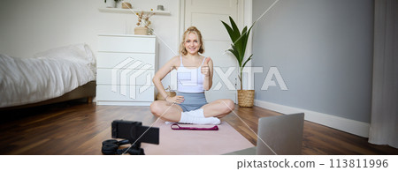 Fitness blogger, woman records video of herself, sitting on rubber mat for exercises in front of digital camera and laptop, shows thumbs up, creates content for social media account about wellbeing 113811996