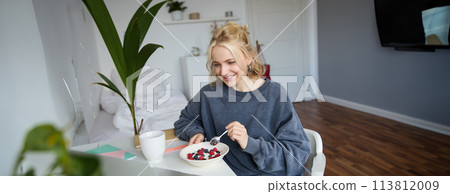 Portrait of smiling blond young woman, eating in front of laptop, watching videos online while having breakfast, enjoying dessert, sitting in bedroom 113812009