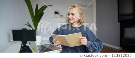 Portrait of cute young woman, teenage girl records video on digital camera, uses laptop to create lifestyle content, shows notebook, reads notes, using computer in her room 113812023