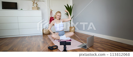 Young female athlete, fitness instructor woman sits on floor rubber mat, recording video on digital camera, showing how to workout, explaining exercises 113812068