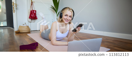 Portrait of young sporty woman, connects to online workout training session, doing exercises, lying on rubber mat, listens to fitness instructor in headphones, holding mobile phone Portrait of young sporty woman, connects to online workout training session, doing exercises, lying on rubber mat, listens to fitness instructor in headphones, holding mobile phone 113812113